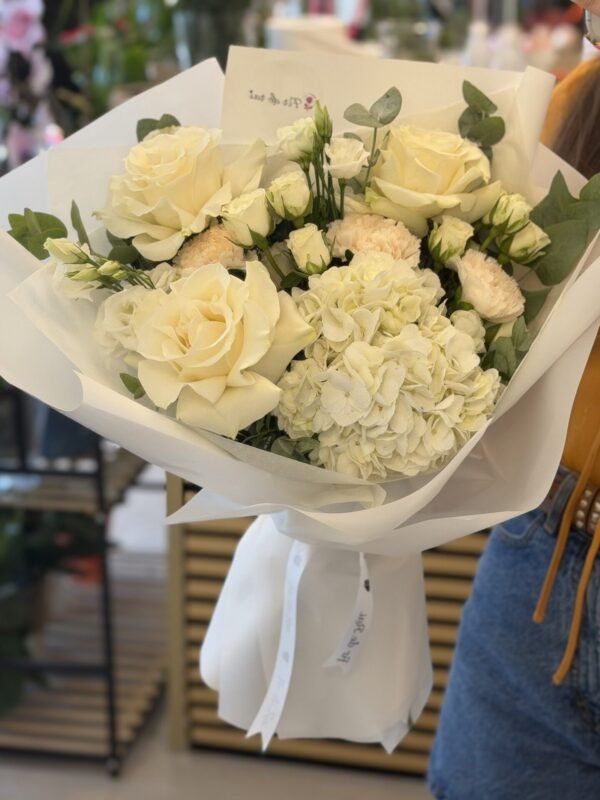 Buchet premium alb cu hortensie, trandafiri ivoire, miniroze și garoafe crem, ambalat elegant în hârtie albă.