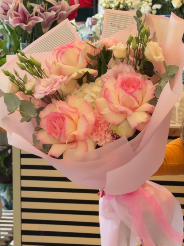 Buchet premium cu trandafiri crem cu margini roz, lisianthus și eucalipt, ambalat elegant în hârtie roz pastel.
